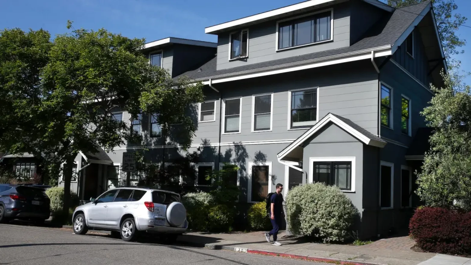 A man in a black shirt and blue jeans, wearing a black backpack and glasses, walks past a gray multifamily housing structure.